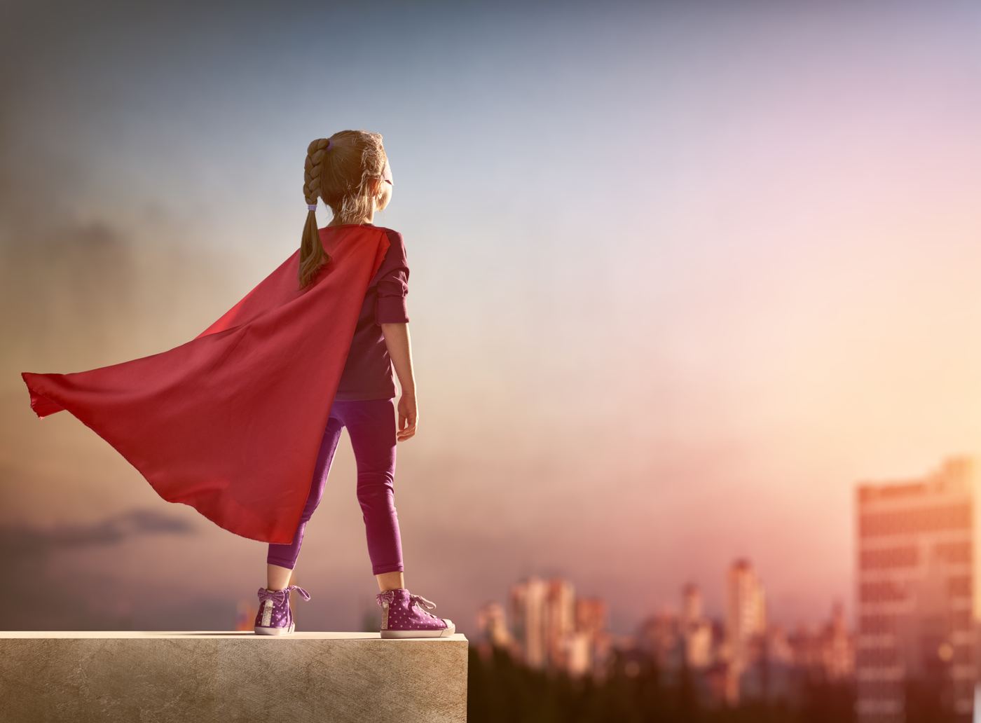 A young girl in a red superhero cape looking out over a city at sunset